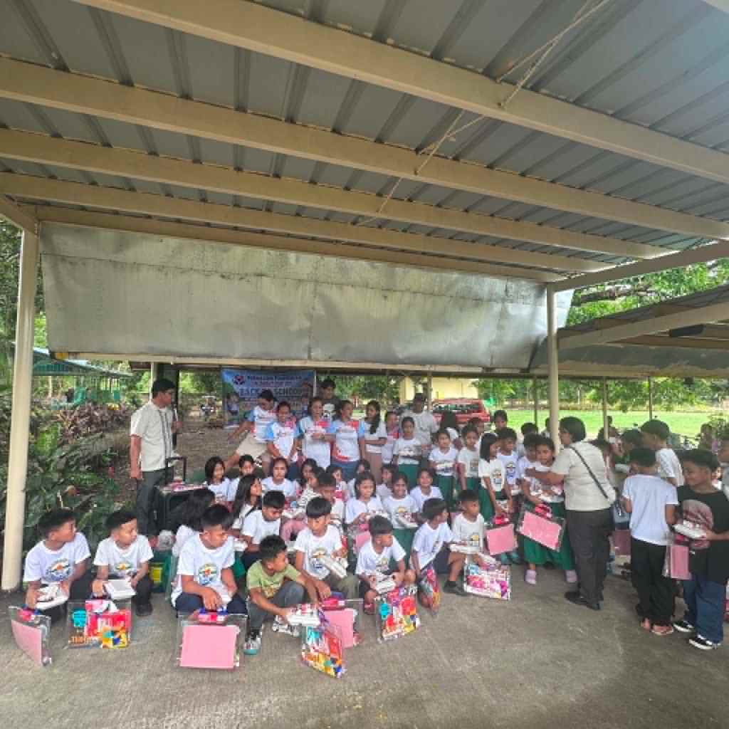 A group of children wearing matching t-shirts gather under a shelter, holding colorful school supplies and smiling. Adults are interacting with them, and a banner is visible in the background.