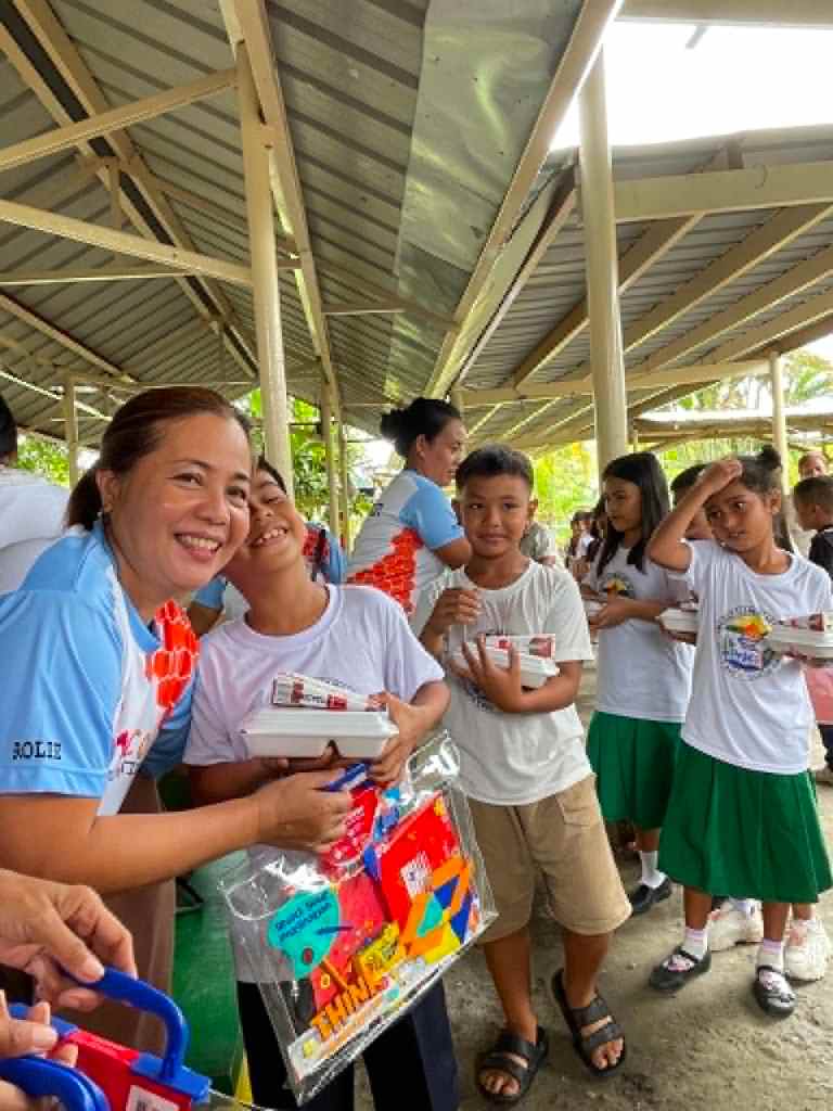 A smiling woman distributing school supplies to children in a covered area, with several kids holding educational materials and toys.