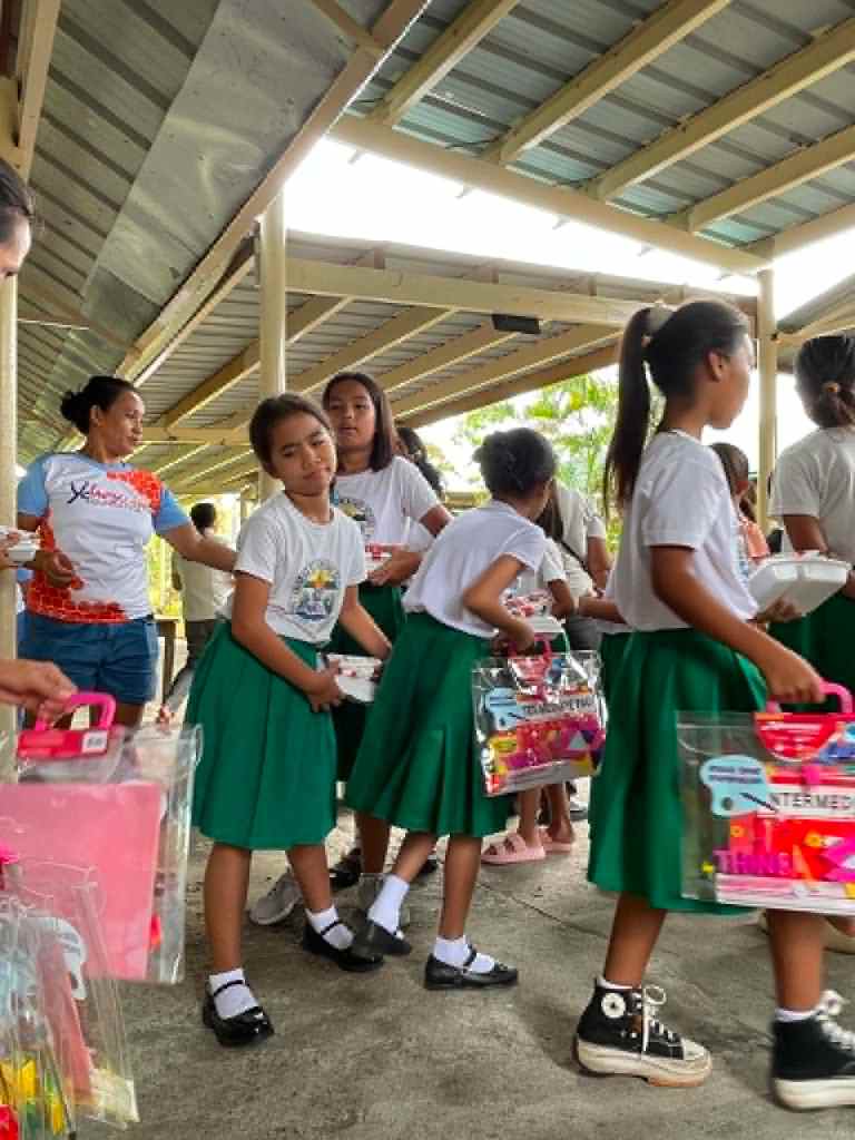 A group of children in green skirts and white shirts carrying clear plastic bags filled with school supplies, standing in a covered outdoor area.