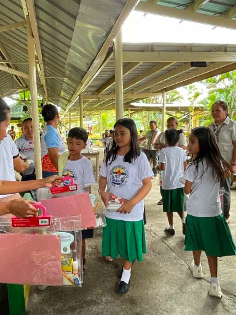 Children in white t-shirts and green skirts line up to receive school supplies outdoors under a sheltered area.