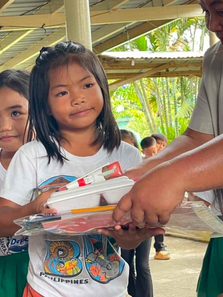 A young girl receiving school supplies from an adult, with another girl standing beside her, in a school setting outdoors.