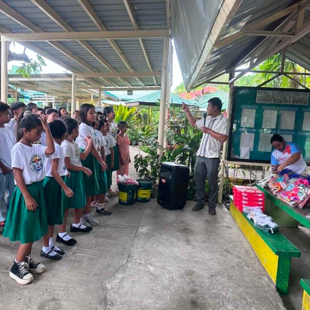 A group of school children in green skirts listening attentively to a man speaking in a covered outdoor area, with plants and a bulletin board in the background.