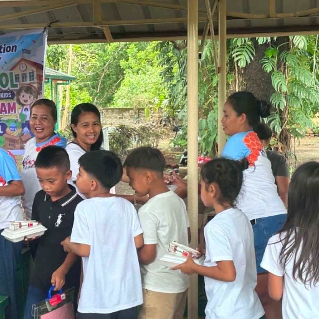 A group of children and adults smiling and interacting at an outdoor event focused on distributing school supplies, with a banner in the background.