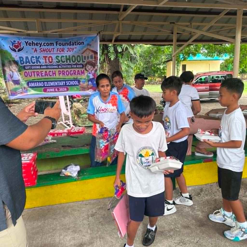 A community outreach event at Amarao Elementary School, featuring a banner for the Yehey.com Foundation's 'Back to School' program. Children and adults are seen participating in gift giving and feeding activities, with several kids holding school supplies.