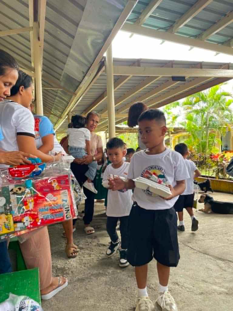Children receiving gifts in a community event, with adults interacting in an outdoor setting.