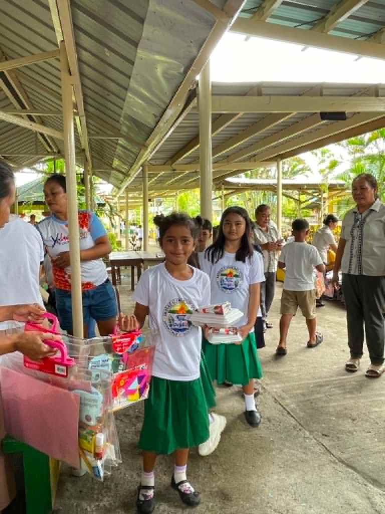 Children in white shirts and green skirts participating in an outdoor event, holding school supplies and snacks while interacting with adults in a communal area.