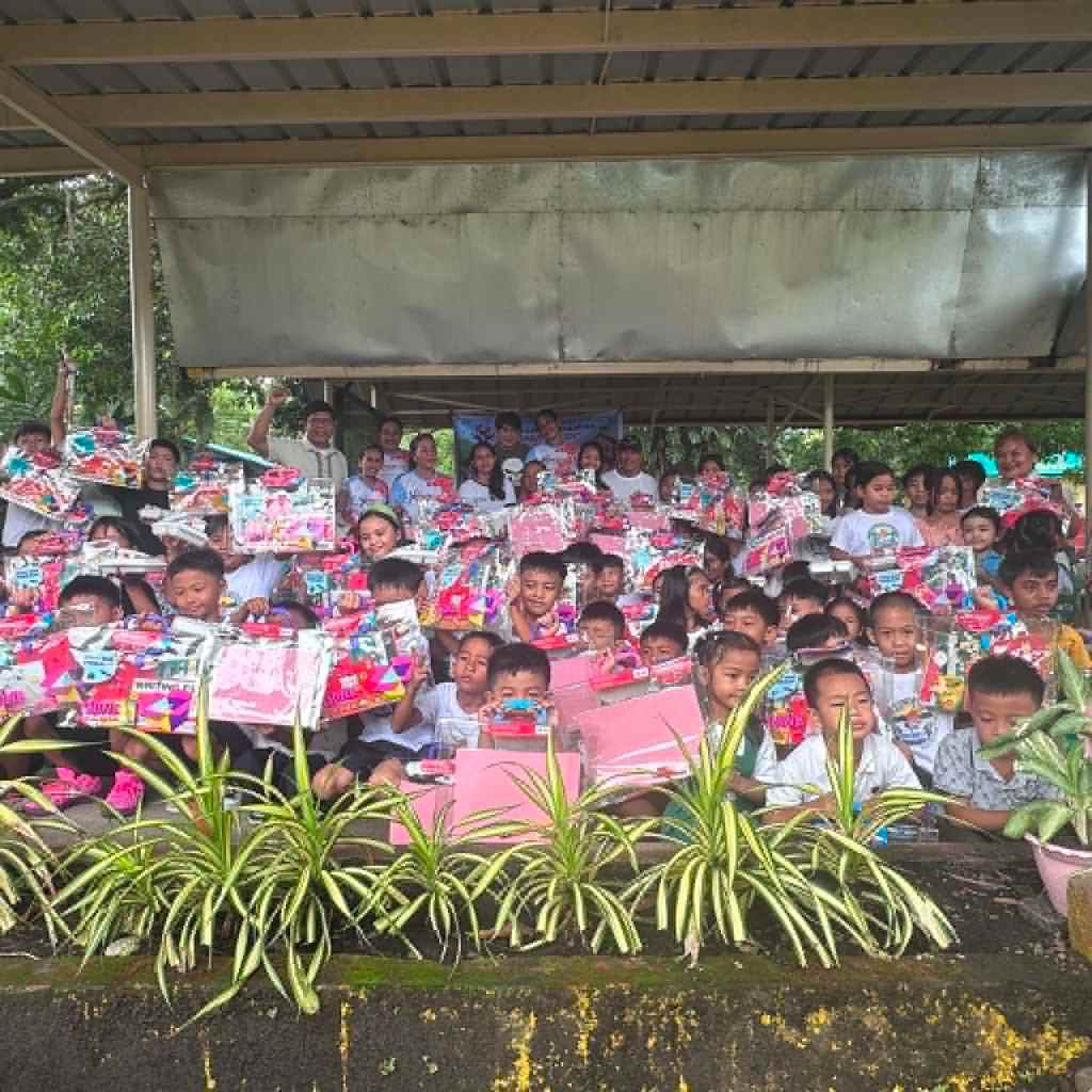 A large group of children and adults gathered under a shelter, holding colorful gift packages and smiling, with greenery in the foreground.