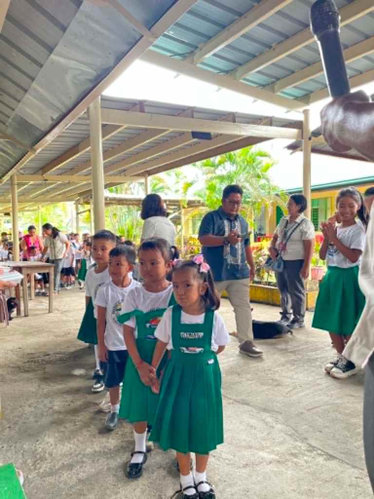 A group of young children in green uniforms waiting in line during an outdoor school event, with adults and other students in the background.