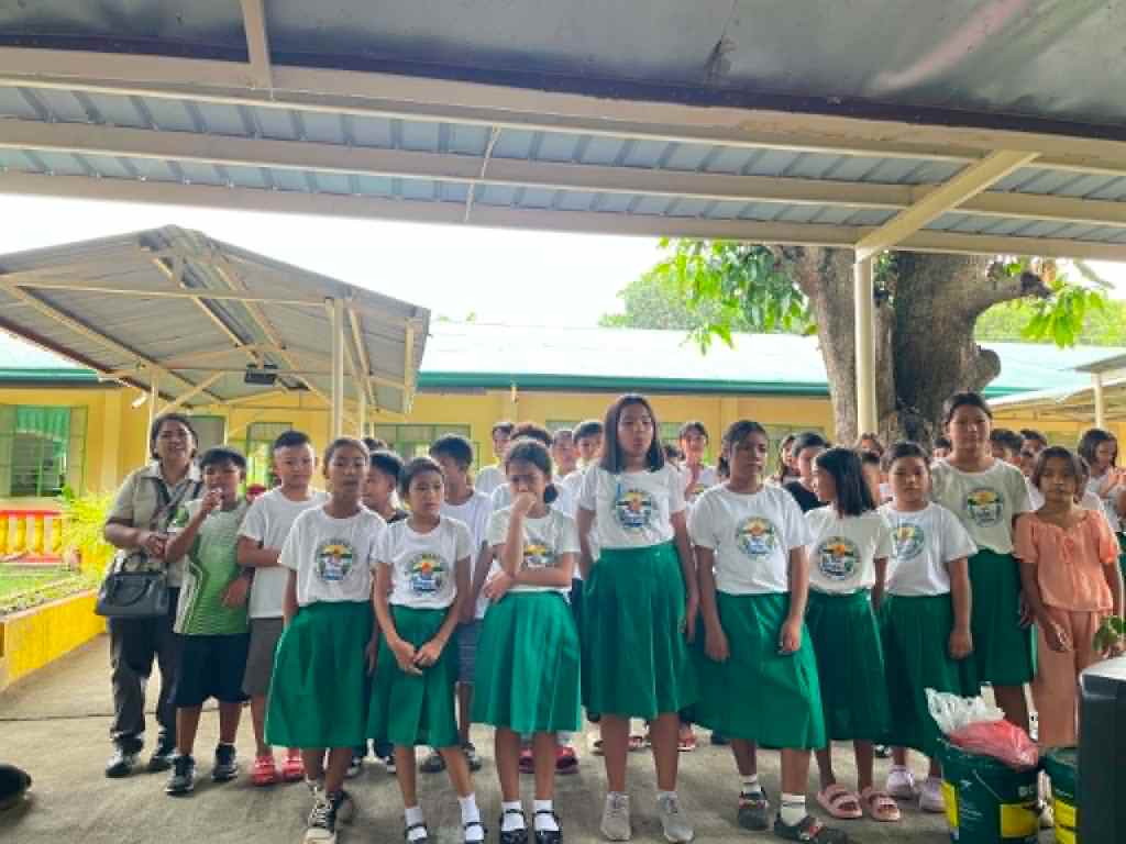 Group of schoolchildren wearing uniforms with white shirts and green skirts, standing together outdoors, with a few adults in the background.