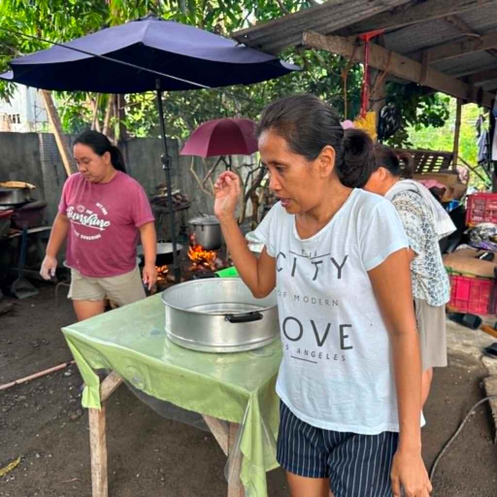 Two women are preparing food at a makeshift outdoor kitchen. One woman, wearing a white shirt with text, is standing next to a pot on a table, while another woman in a pink shirt is visible in the background.