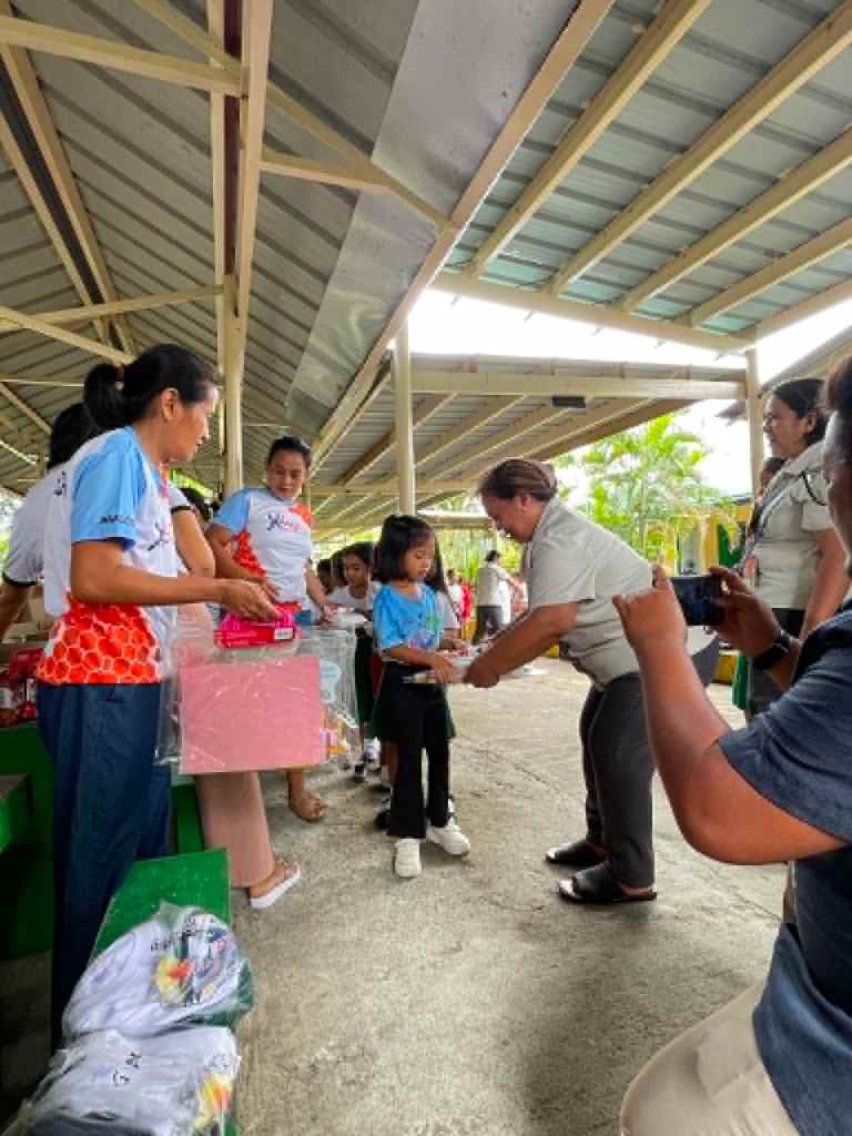 A group of adults and children participating in a community event where supplies or gifts are being distributed, with several people actively engaged in handing out items under a covered area.