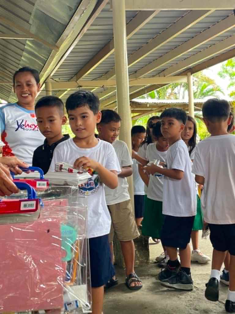 A group of children in white t-shirts lining up to receive school supplies from a smiling adult, under a sheltered area.