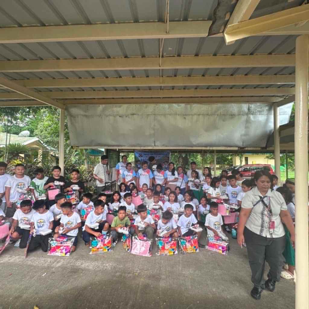 Group photo of children and adults at an outdoor event, some holding colorful gift bags, under a covered area.