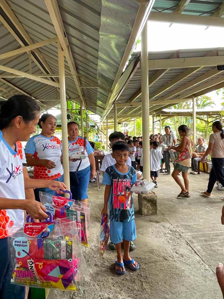 A group of adults in matching shirts are handing out colorful gift bags to children in a covered outdoor area.