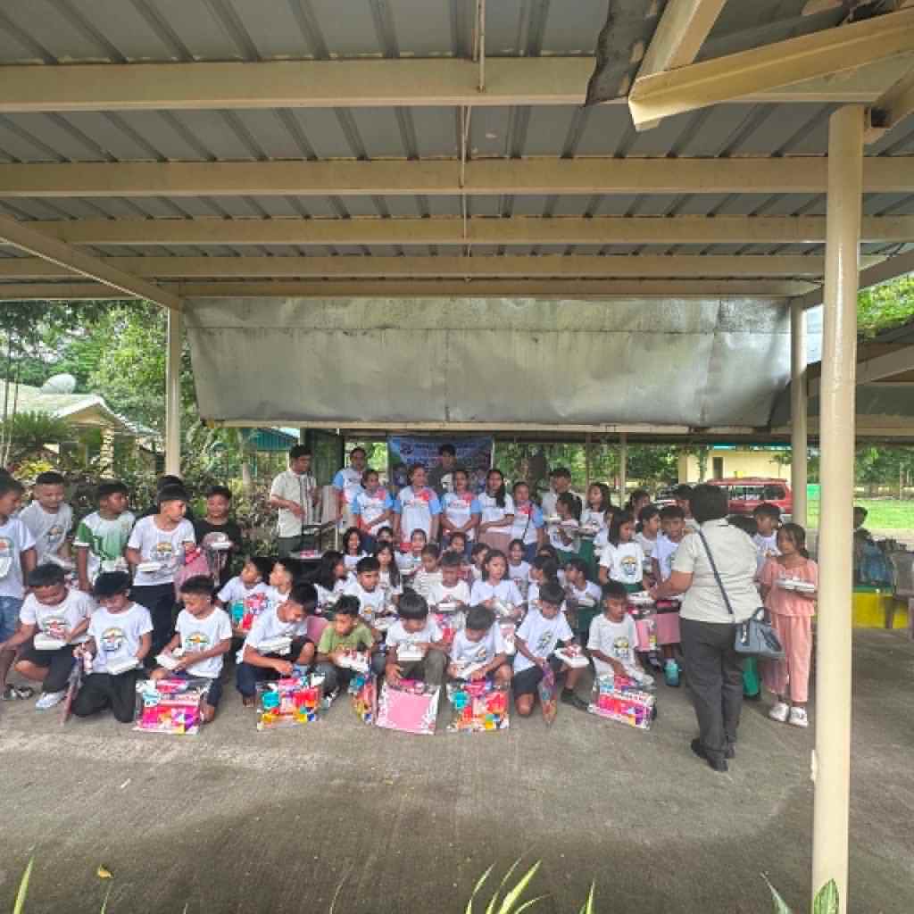 A group of children wearing white T-shirts, gathered under a shelter, holding gifts and posing for a photo during a community event.