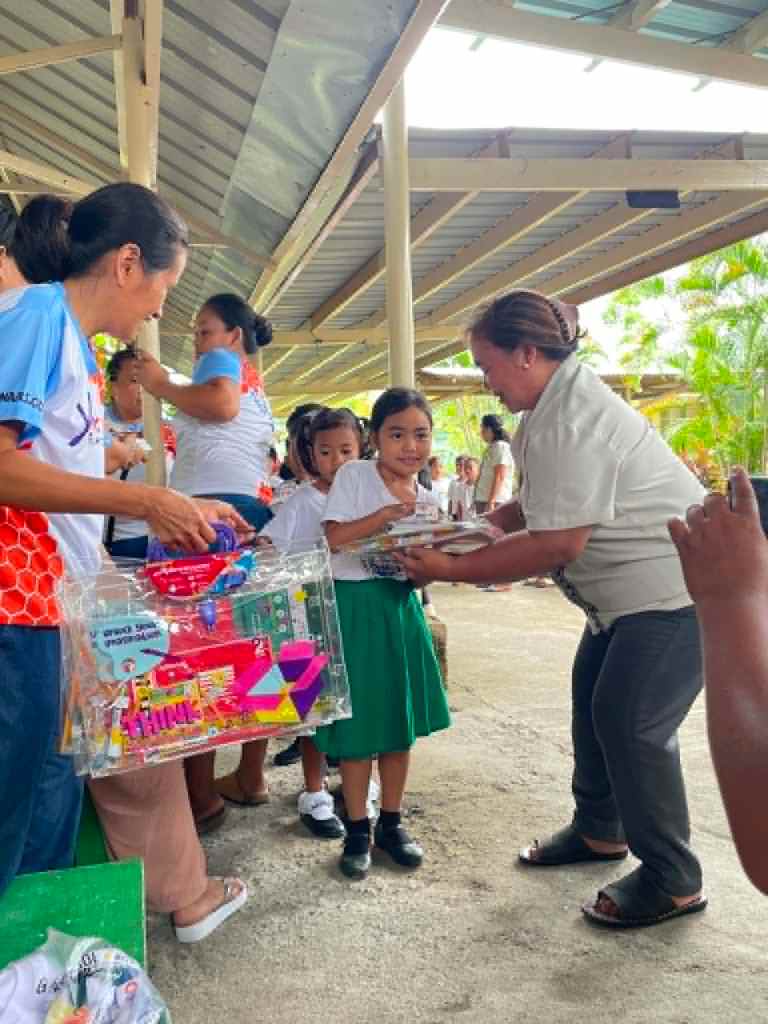 Children receiving supplies at an outdoor distribution event, with adults handing out items in a school setting.