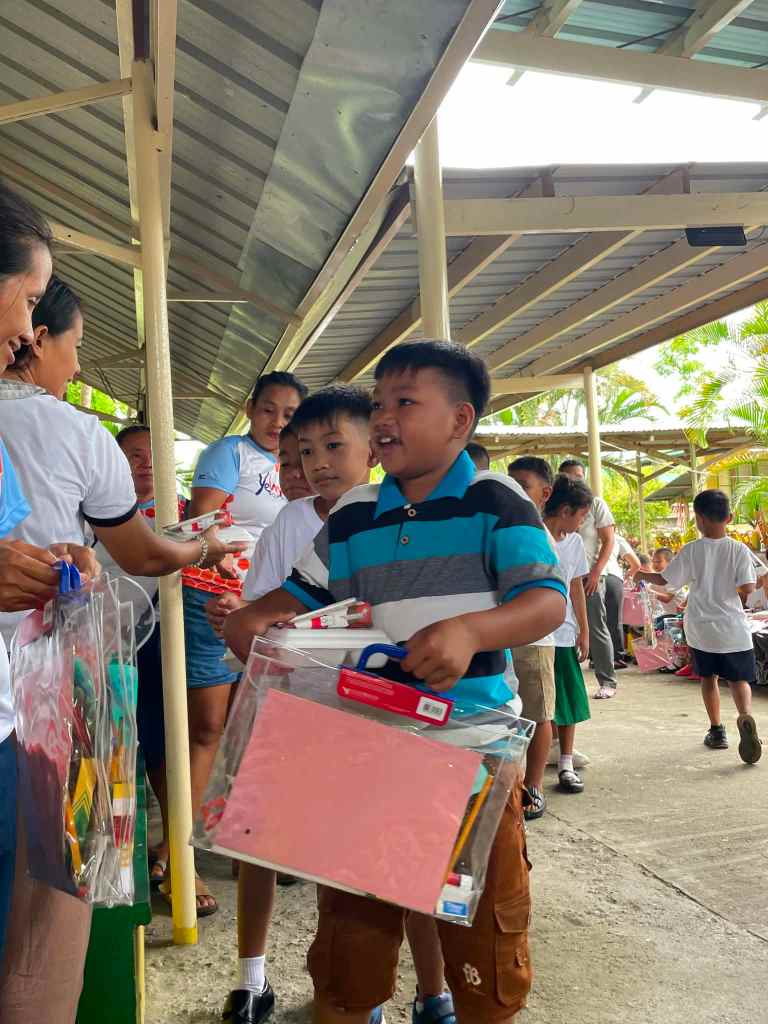 Children receiving school supplies in clear bags during an event, smiling as they interact with adults in a covered outdoor setting.