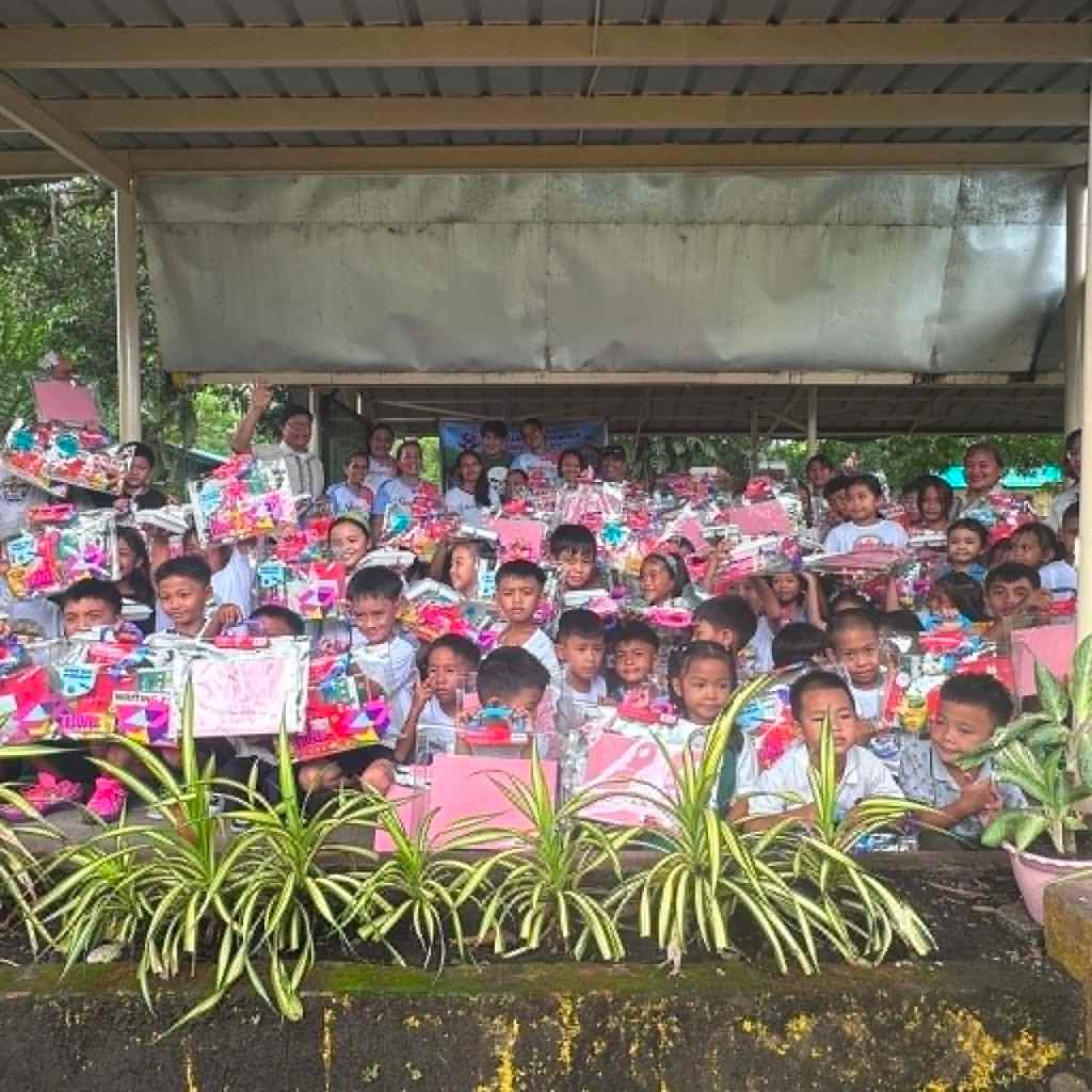 A large group of children and adults, smiling and holding colorful gift packages, gathered under a shelter surrounded by greenery.