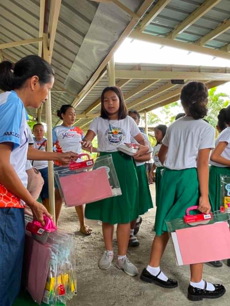 A group of girls in green skirts and white shirts receiving colorful school supplies from a woman in a blue shirt under a covered area.
