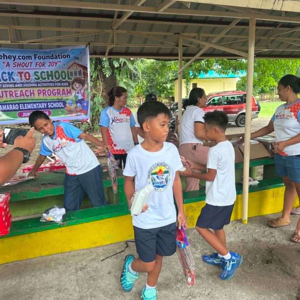 A community outreach program at a school with children receiving supplies and volunteers helping in the background.